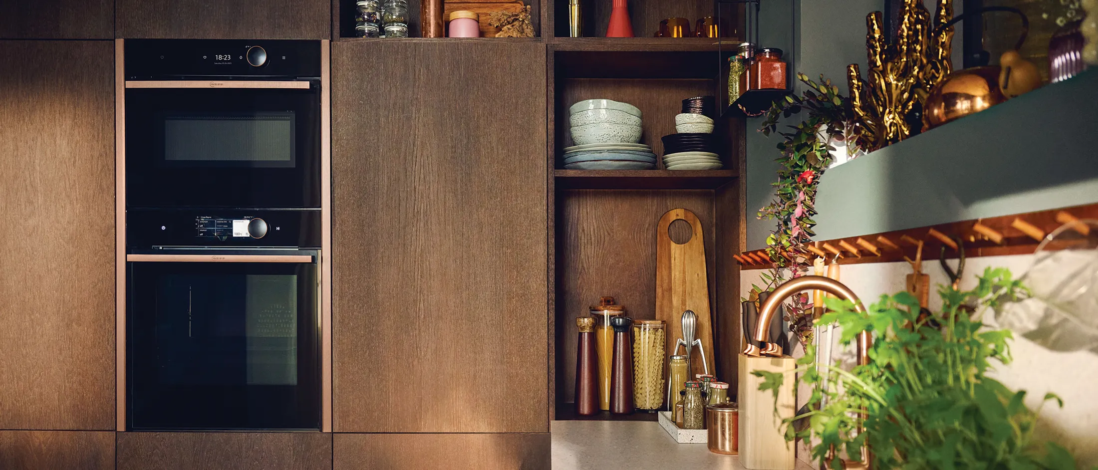 Moderne Küchenecke mit Einbaubacköfen, Holzregalen mit Geschirr, Gewürzen und Schneidebrettern sowie einer Spüle mit Grünpflanzen in der Nähe.