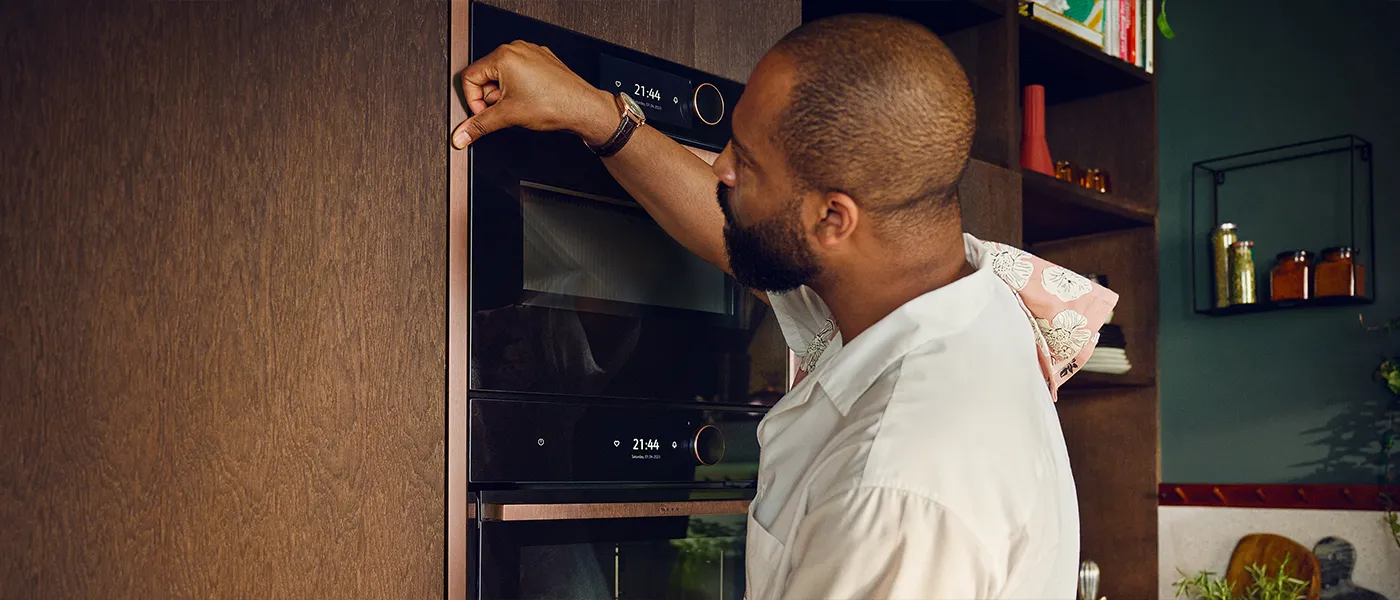 Uomo con una camicia bianca che estrae da un forno aperto una teglia di pollo arrosto con verdure, in cucina.