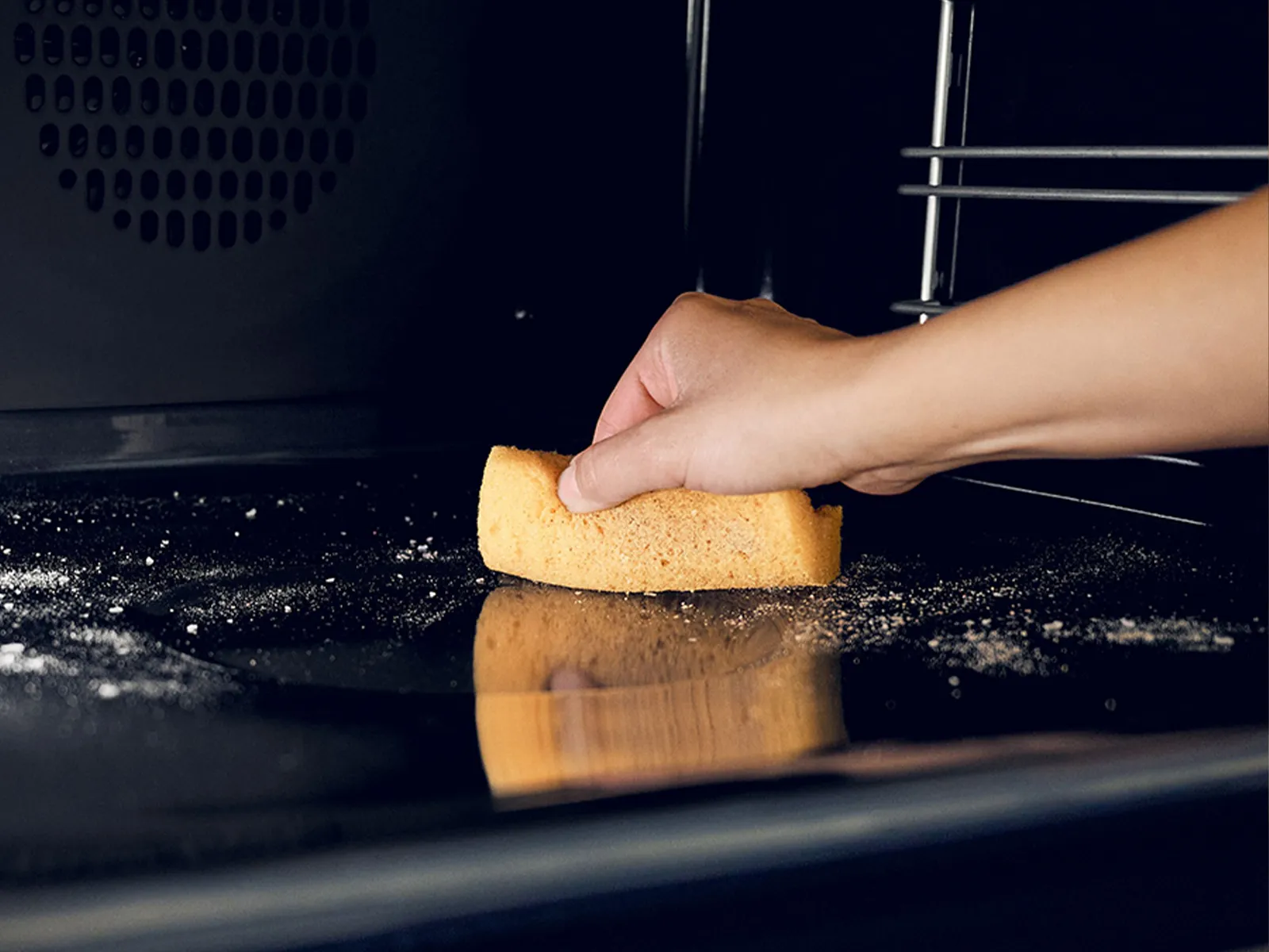 Χέρι καθαρίζει το εσωτερικό ενός φούρνου με κίτρινο σφουγγάρι, απομακρύνοντας μαύρα κατάλοιπα και βρομιά από τις επιφάνειές του.