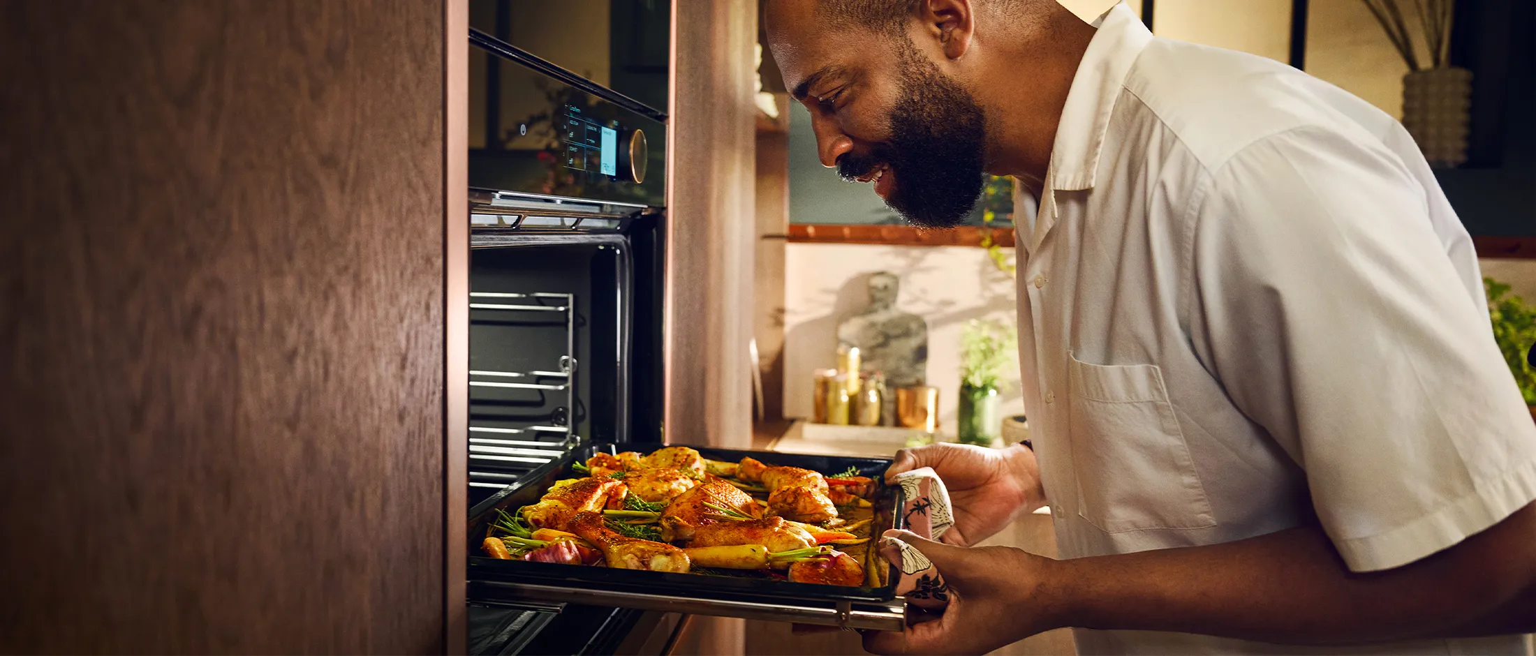 Man wearing a white shirt removes a tray of roasted chicken with vegetables from an open oven in a kitchen.
