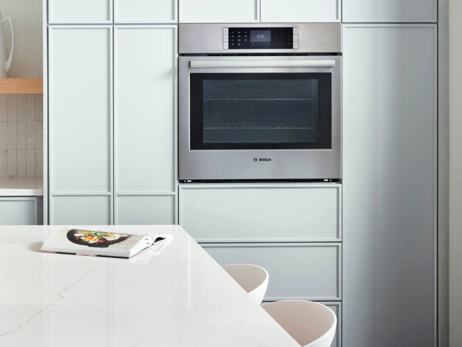 Cuisine blanche moderne avec une cuisinière à gaz, un four encastré, une crédence minimaliste et un îlot épuré avec des tabourets et une assiette posée dessus.