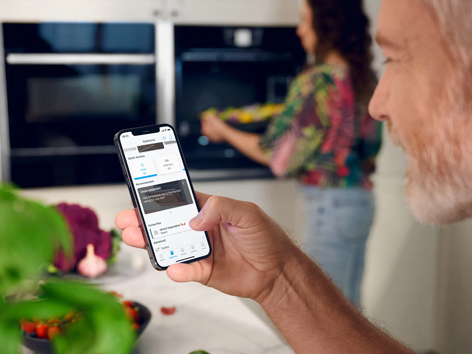 Man holds smartphone, woman stands by the oven in the background