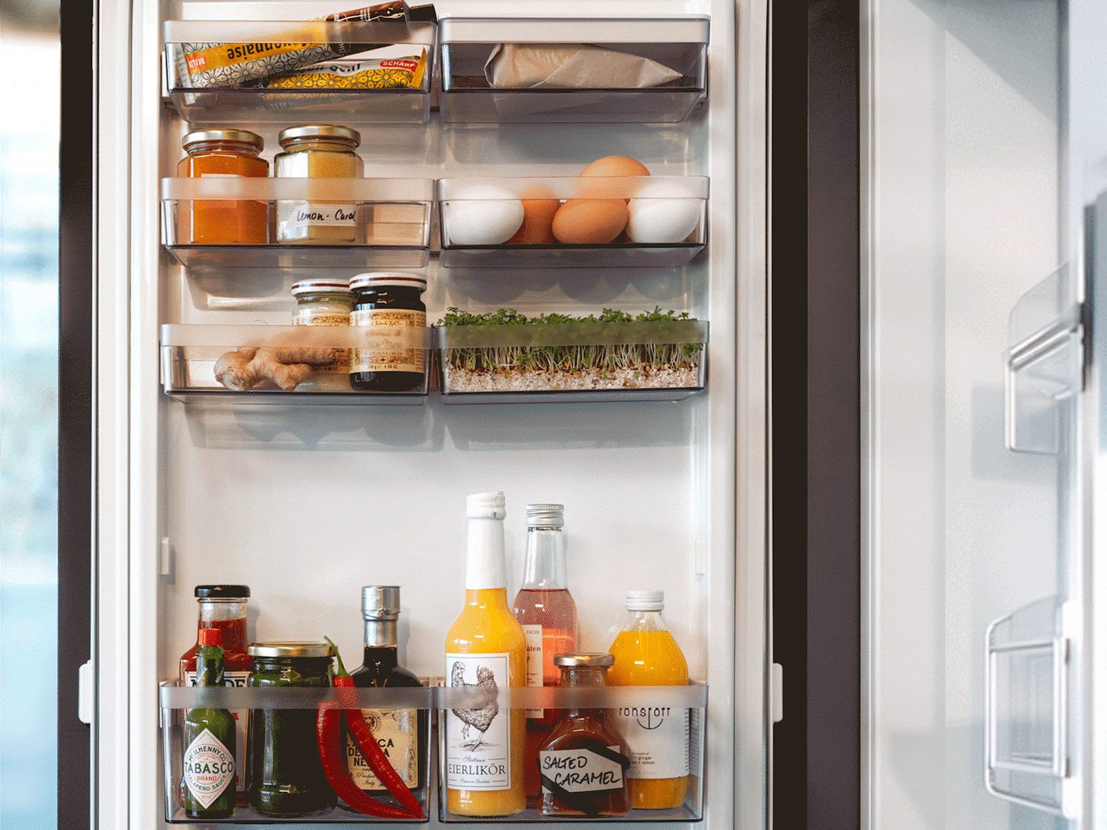 Ein gut organisierter Kühlschrank in dem farbenfrohe Lebensmittel mit Einmachgläsern, Eiern, frischen Kräutern, verschiedenen Flaschen und Milch gut sichtbar sind.