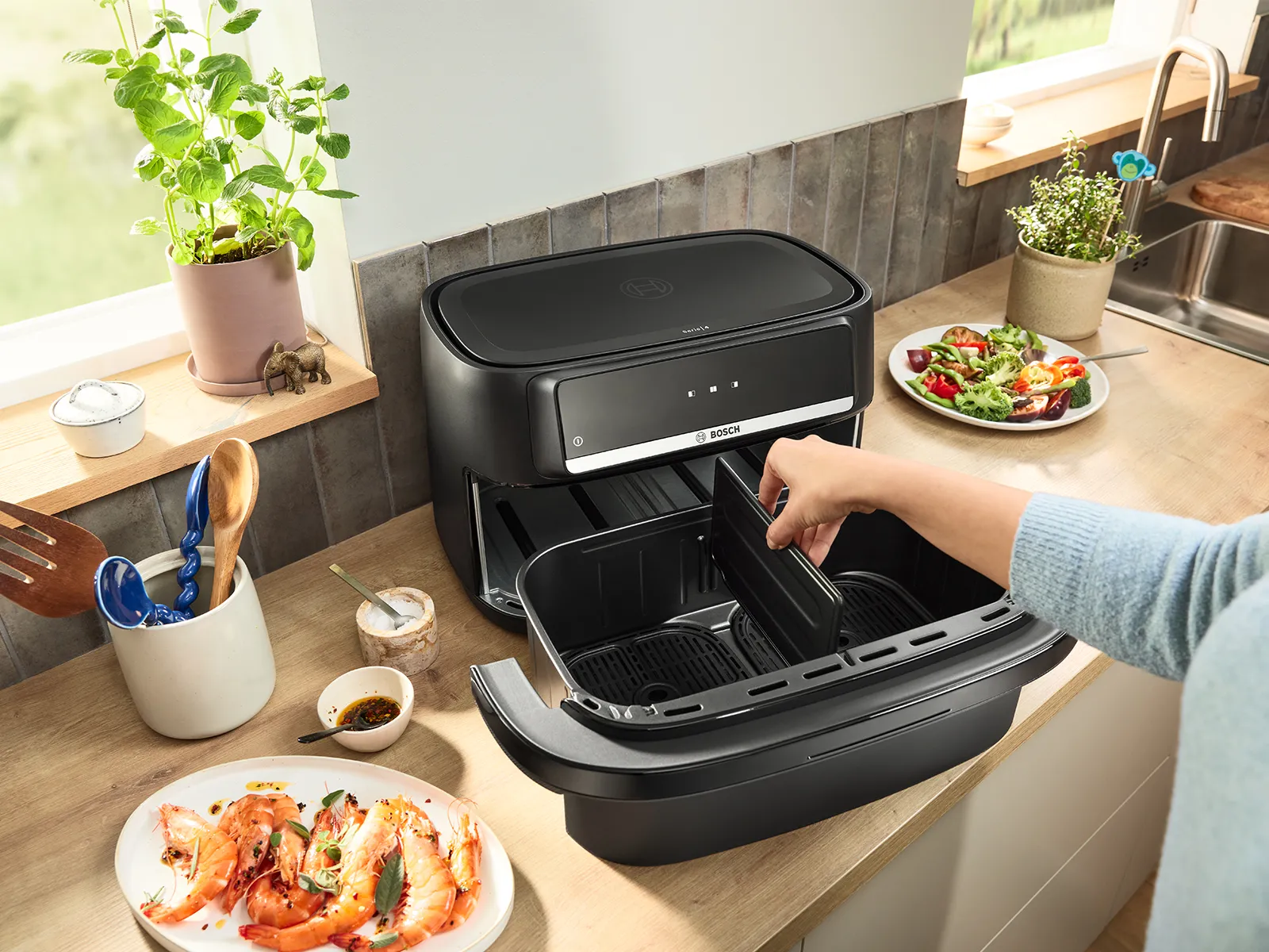 A person places a divider into a black Bosch air fryer with dual baskets on a wooden kitchen counter. Plates of cooked shrimp and fresh salad are nearby.