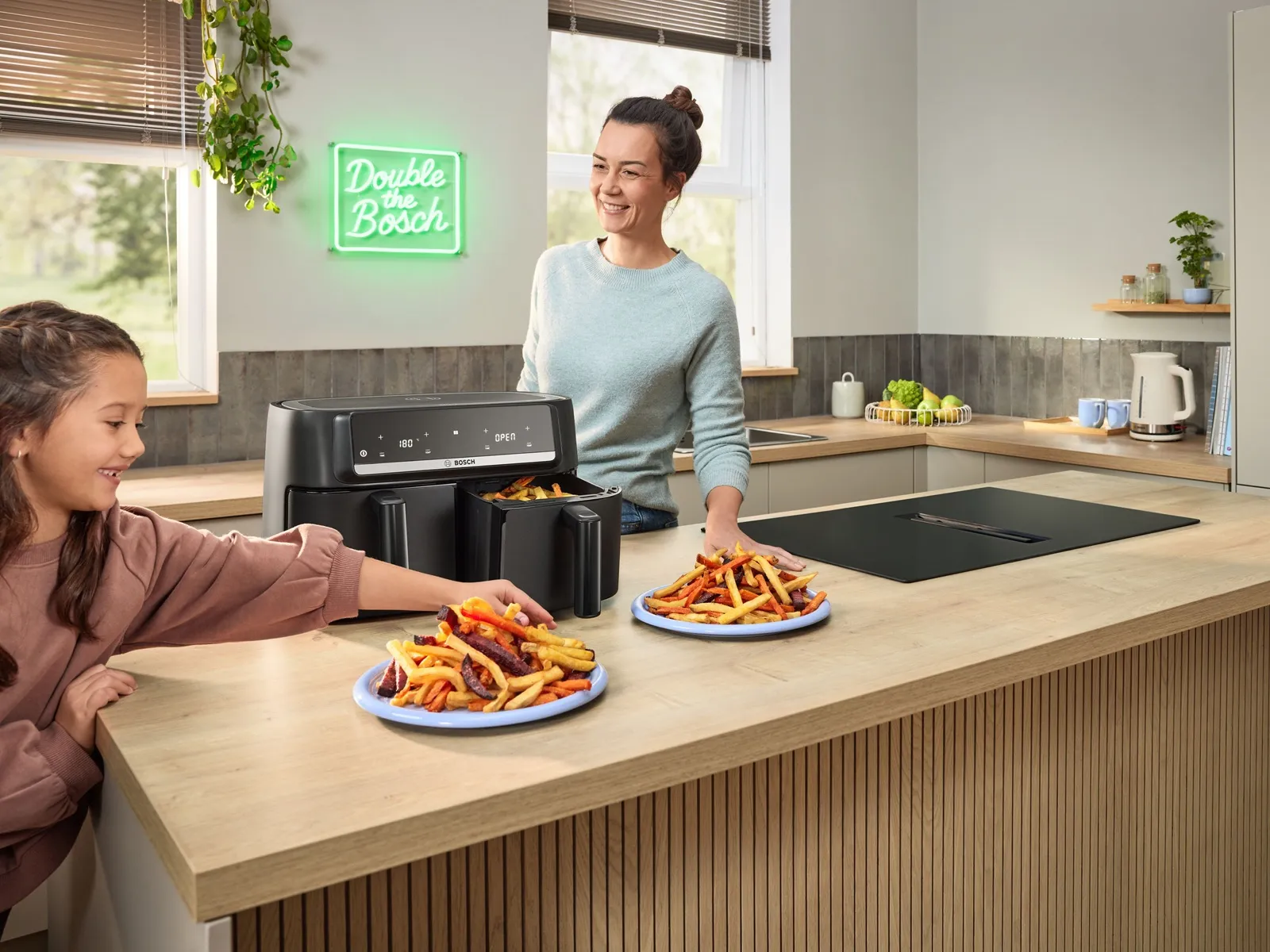 A Bosch Double Drawer Air Fryer is open on the kitchen side with colourful chips in, a woman and a child take chips off the two plates in front.