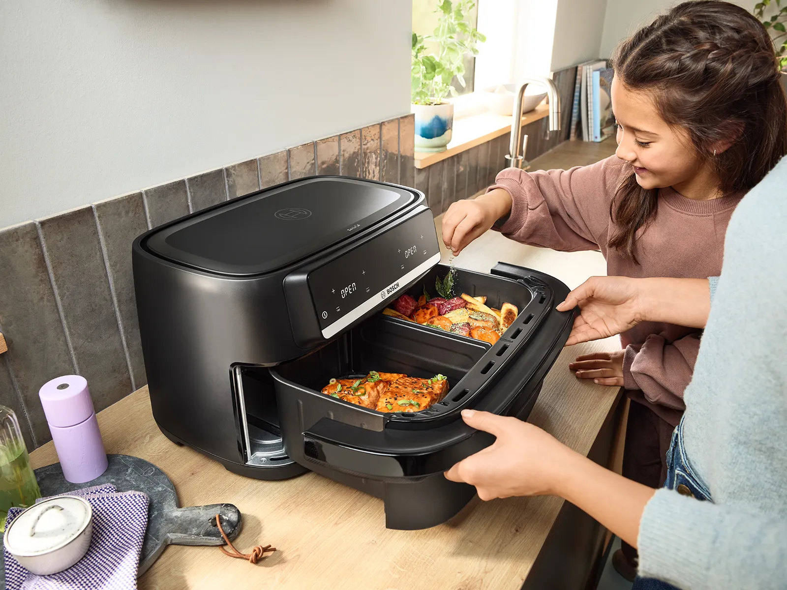 A woman opening the Bosch Flex Drawer Air Fryer and a small child sprinkling some spice on the food inside.