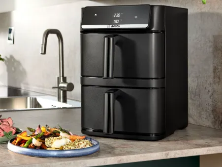 Black Bosch air fryer with digital display on kitchen countertop beside a plate of cooked vegetables and fish.