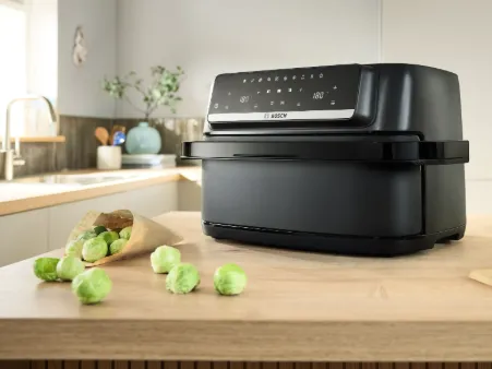 Black Bosch air fryer on a wooden kitchen counter with Brussels sprouts spilled from a paper bag nearby.
