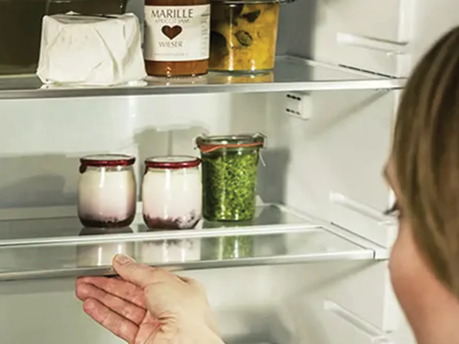 Eine Person greift in einen Kühlschrank, auf dessen Ablagen Joghurtgläser, frische Kräuter und eine Flasche Marillenlikör zu sehen sind.