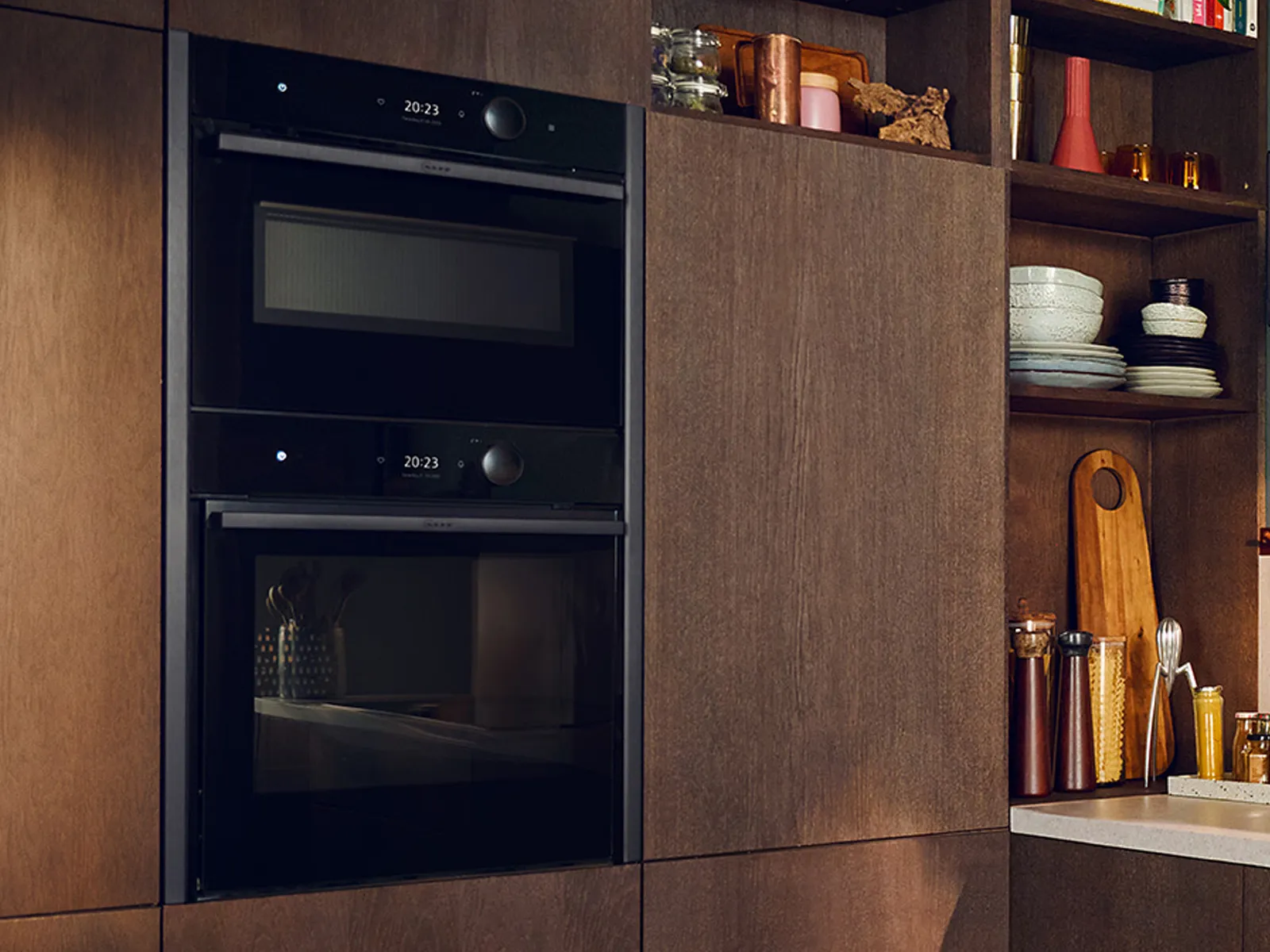 Modern kitchen with built-in black double oven, dark wood cabinetry, open shelves holding dishes, glassware, and kitchen accessories.