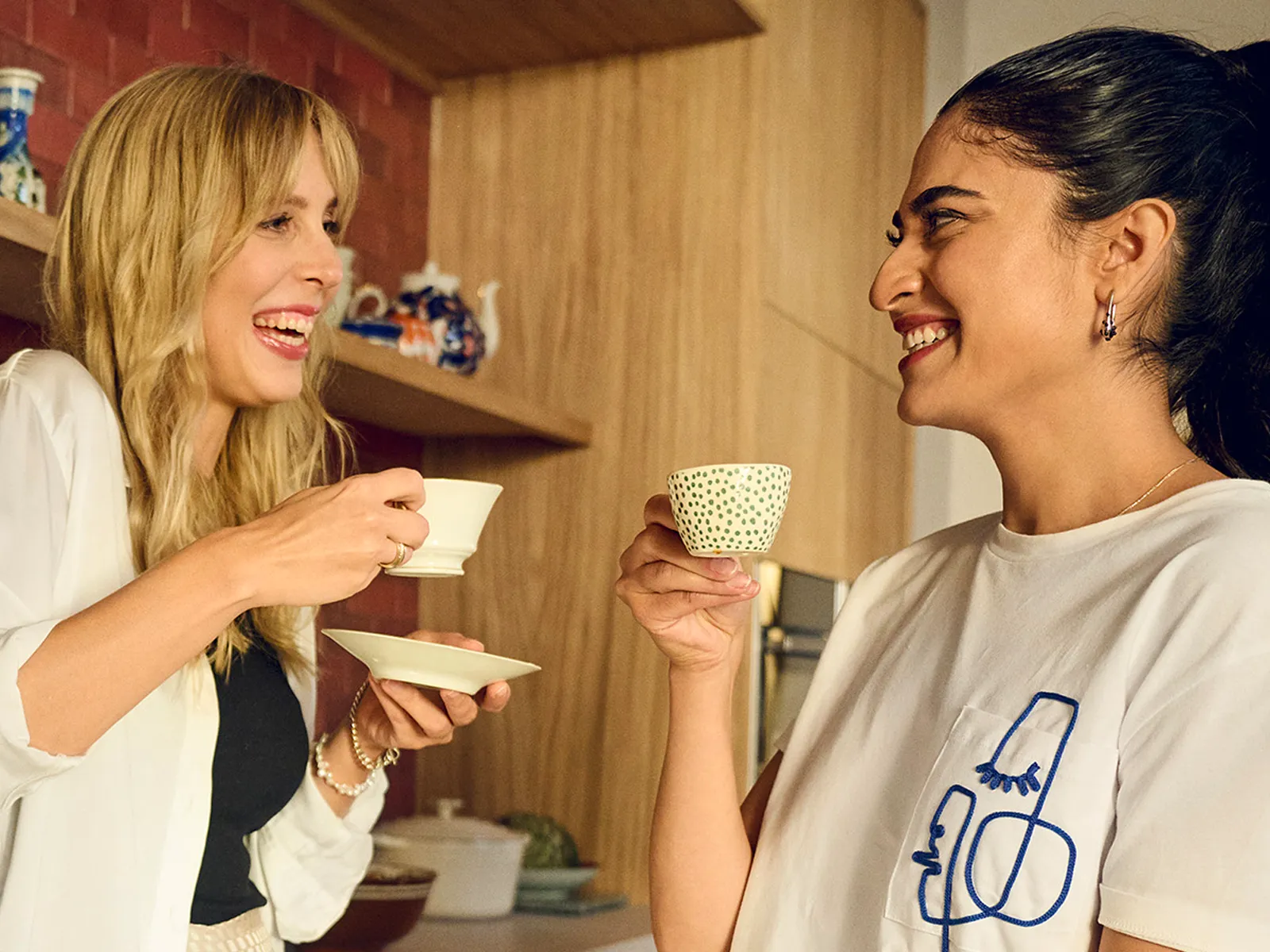 Two women laugh and talk while enjoying a cup of tea together in a modern, stylish kitchen.