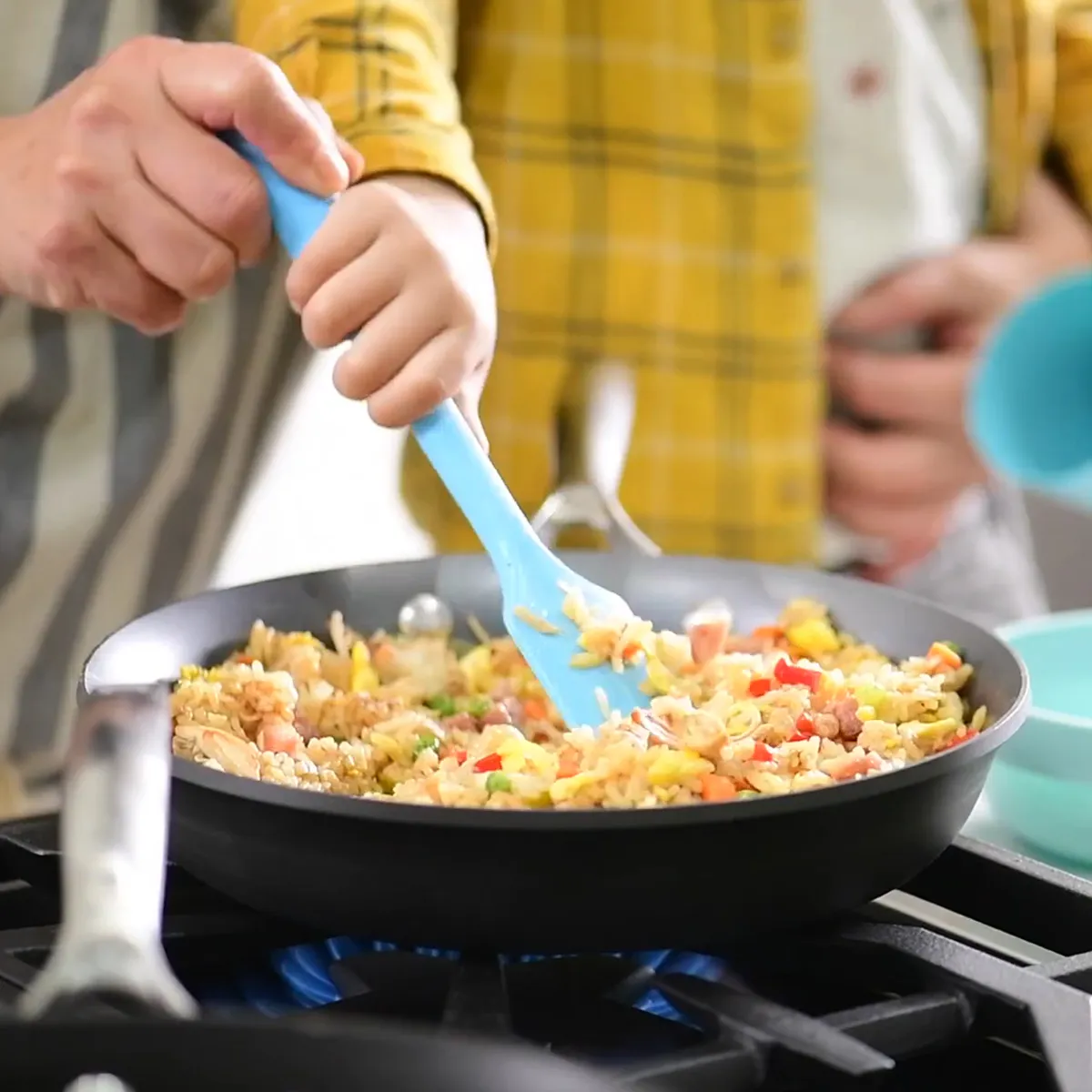 Pan frying a colorful mix of rice, eggs, vegetables, and ham in a skillet with a blue spatula on a stovetop.