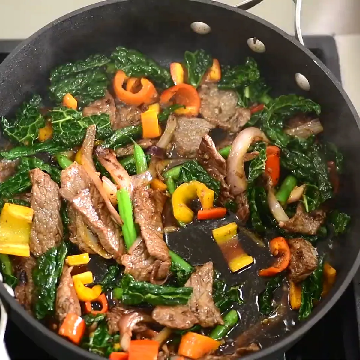 Soy sauce being poured into a pan with stir-fried beef, kale, bell peppers, onions, and green beans on a stovetop.