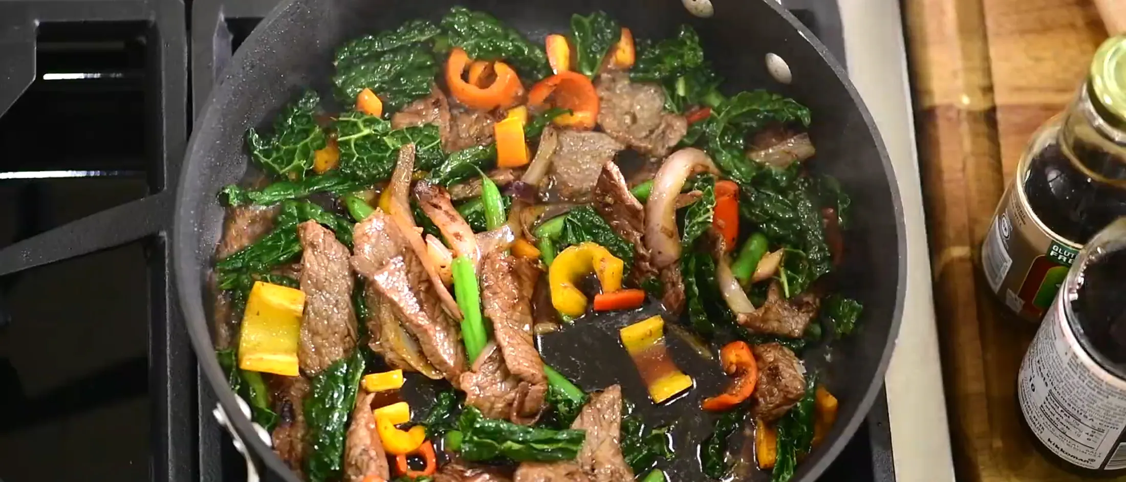 Soy sauce being poured into a pan with stir-fried beef, kale, bell peppers, onions, and green beans on a stovetop.
