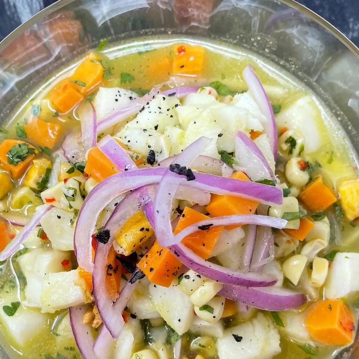 Clear bowl of ceviche with diced fish, orange sweet potato, purple onion slices, corn, and herbs in a light citrus marinade.