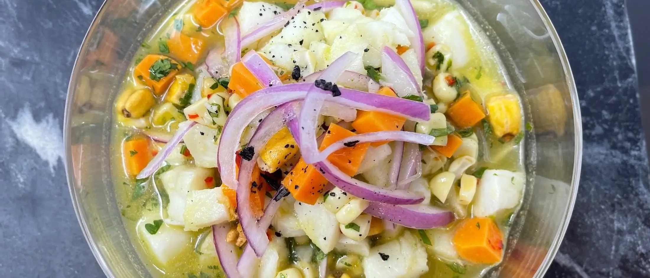 Clear bowl of ceviche with diced fish, orange sweet potato, purple onion slices, corn, and herbs in a light citrus marinade.