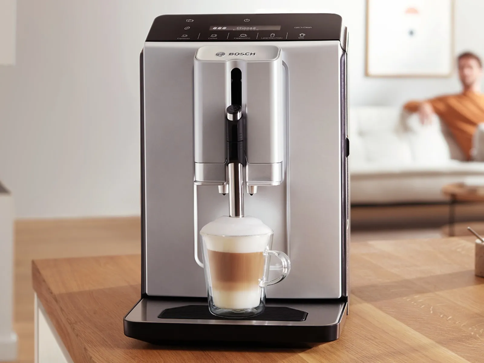 Series 2 VeroCafe coffee machine with cappuccino on kitchen counter. A man sits on the sofa in the background.