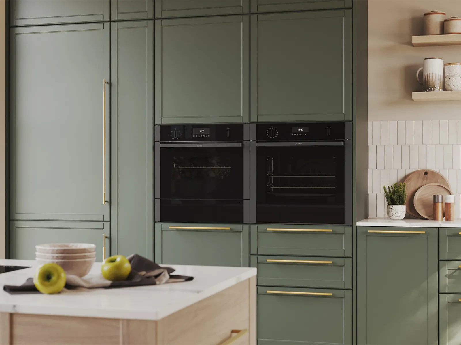 Sage-green kitchen cabinets with brass handles, two NEFF built-in ovens, a white island with bowls and green apples, marble countertop, tiled backsplash and wooden cutting boards.