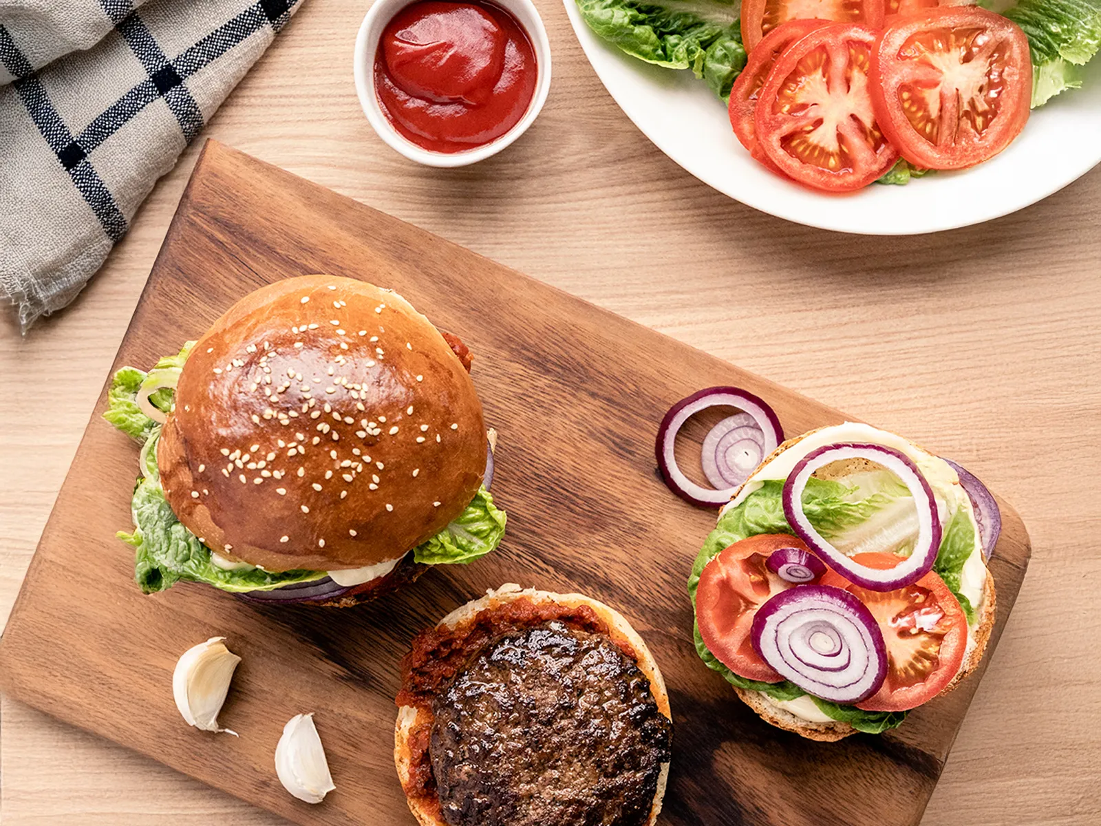 Taxta səthin üzərindən yuxarıdan görünüş: brioche bulkası üzərində burger kotleti, yanında dilimlənmiş pomidor, soğan və kahı yarpaqları.