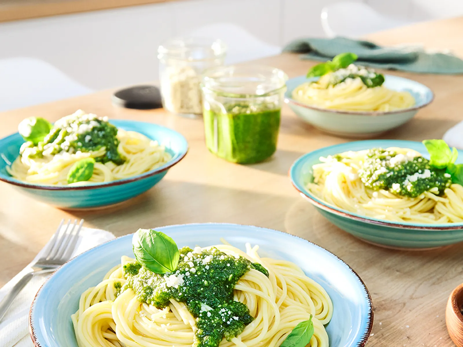 Mərkəzdə pesto dolu banka ilə mətbəx masasının üzərində yaşıl pestolu dörd boşqab makaron düzülüb.