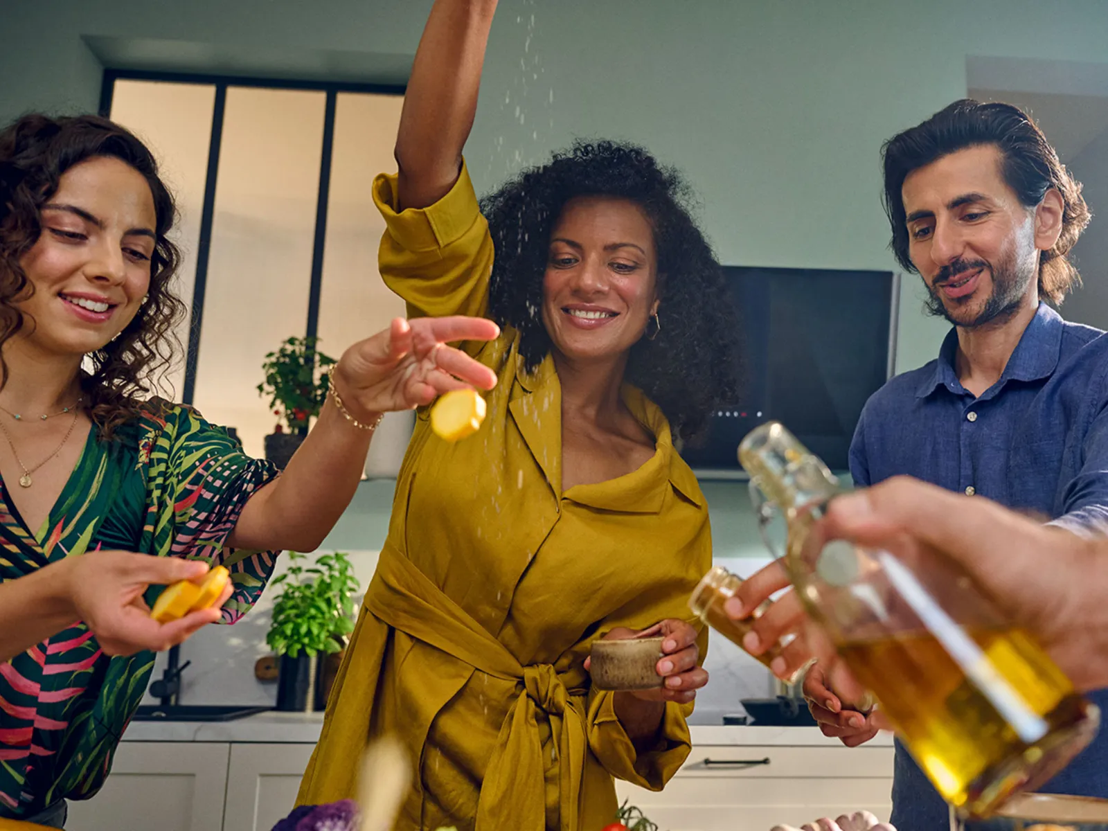 Three friends smiling whilst gathering around an island in a kitchen, one squeezing lemon and another pouring oil over a dish.