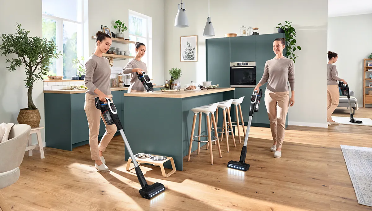 Woman uses Unlimited 9 stick vacuum in a kitchen: cleaning around pet bowls, wiping counters handheld, and walking with the vacuum in a busy home.