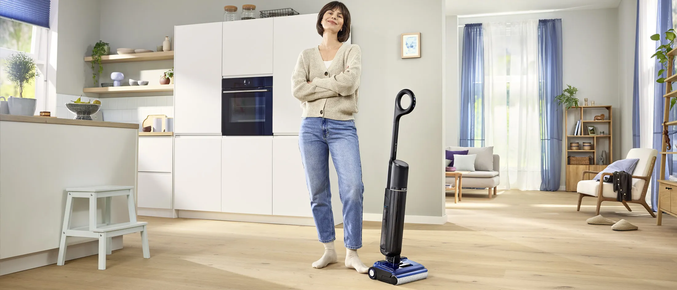 Femme souriante, bras croisés, debout dans une cuisine moderne ouverte sur le salon, à côté d’un aspirateur-balai vertical.