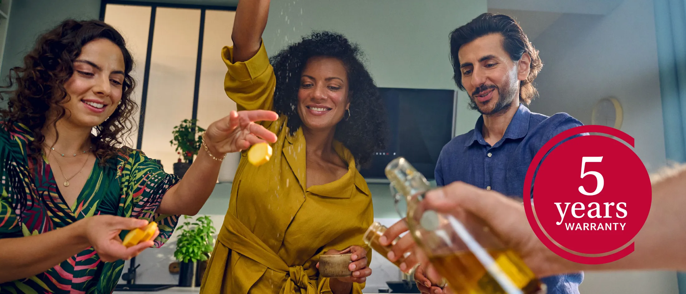 Three people cooking in a modern kitchen: a person in a yellow dress sprinkles seasoning over food, another holds lemon slices, a third watches as a hand in the foreground pours oil; a red circular badge on the right reads "5 years Warranty."