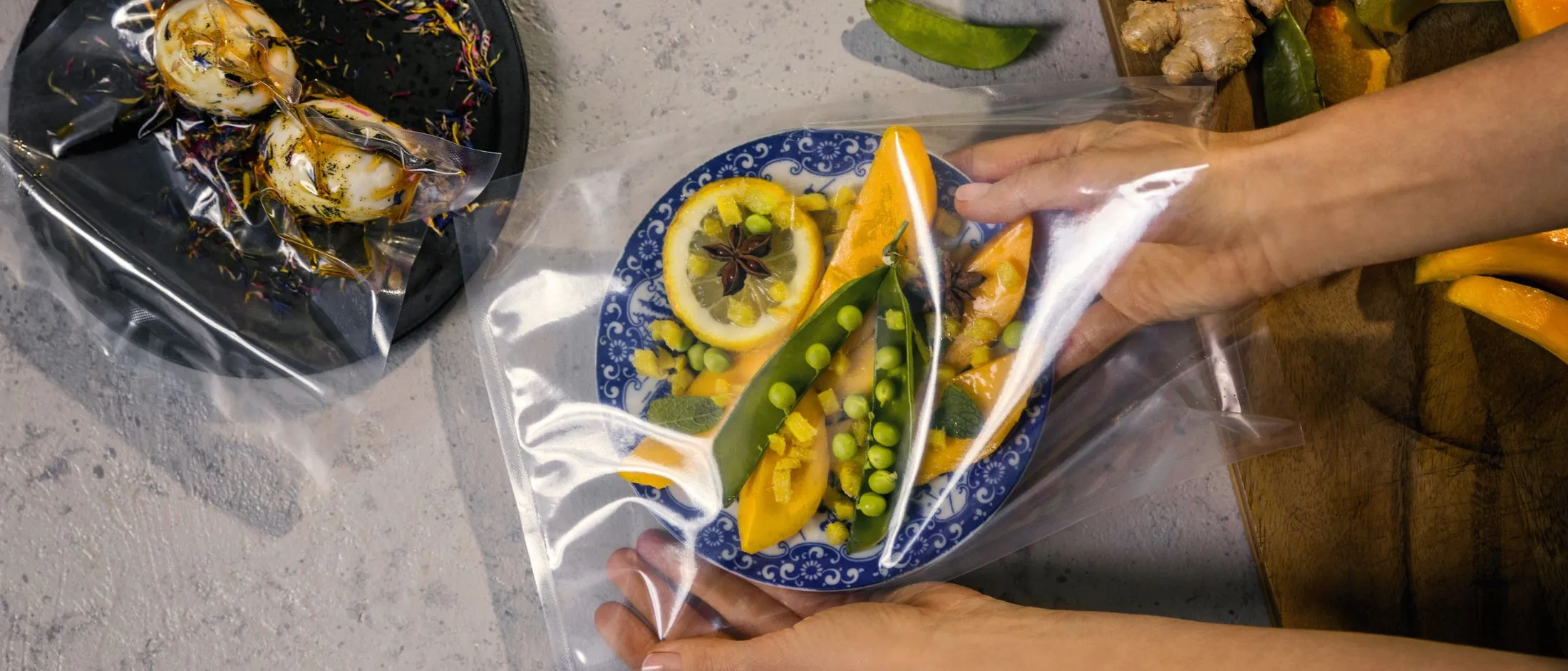 Main posant un film plastique sur une assiette bleue contenant un filet de poisson, quartiers de citron et légumes verts ; à côté, une assiette noire de crustacés.