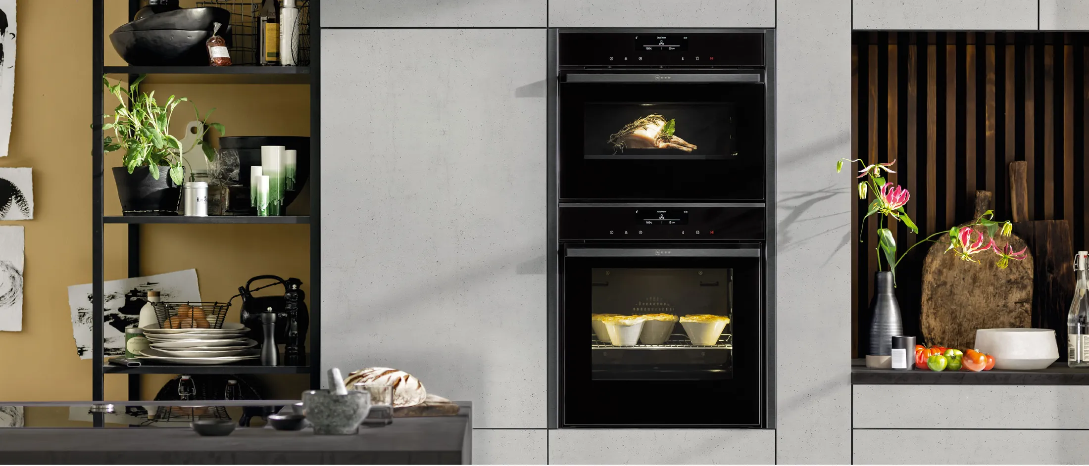 Cucina moderna con forno doppio incorporato, ripiani aperti con piatti e piante e piano di lavoro decorato con fiori e ingredienti da cucina.