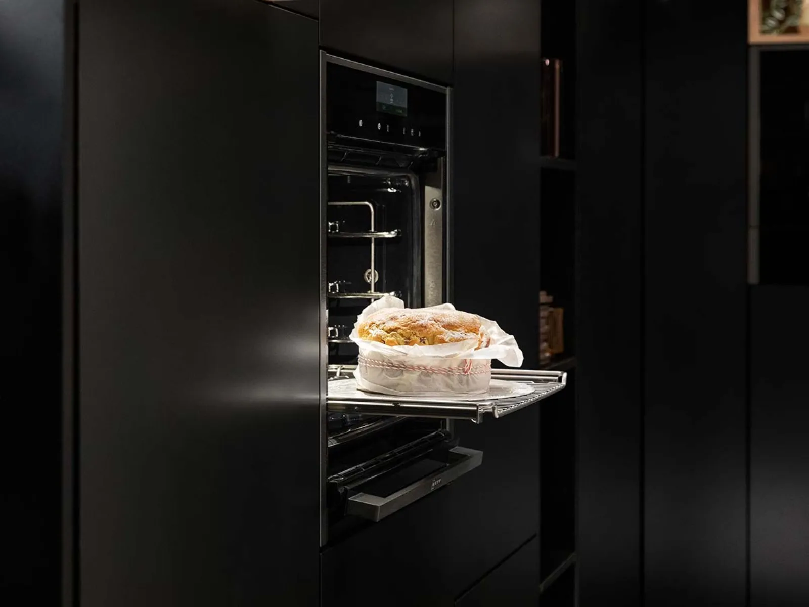 Forno nero aperto con una torta rotonda appena sfornata sulla griglia, circondata da moderni mobili da cucina scuri.
