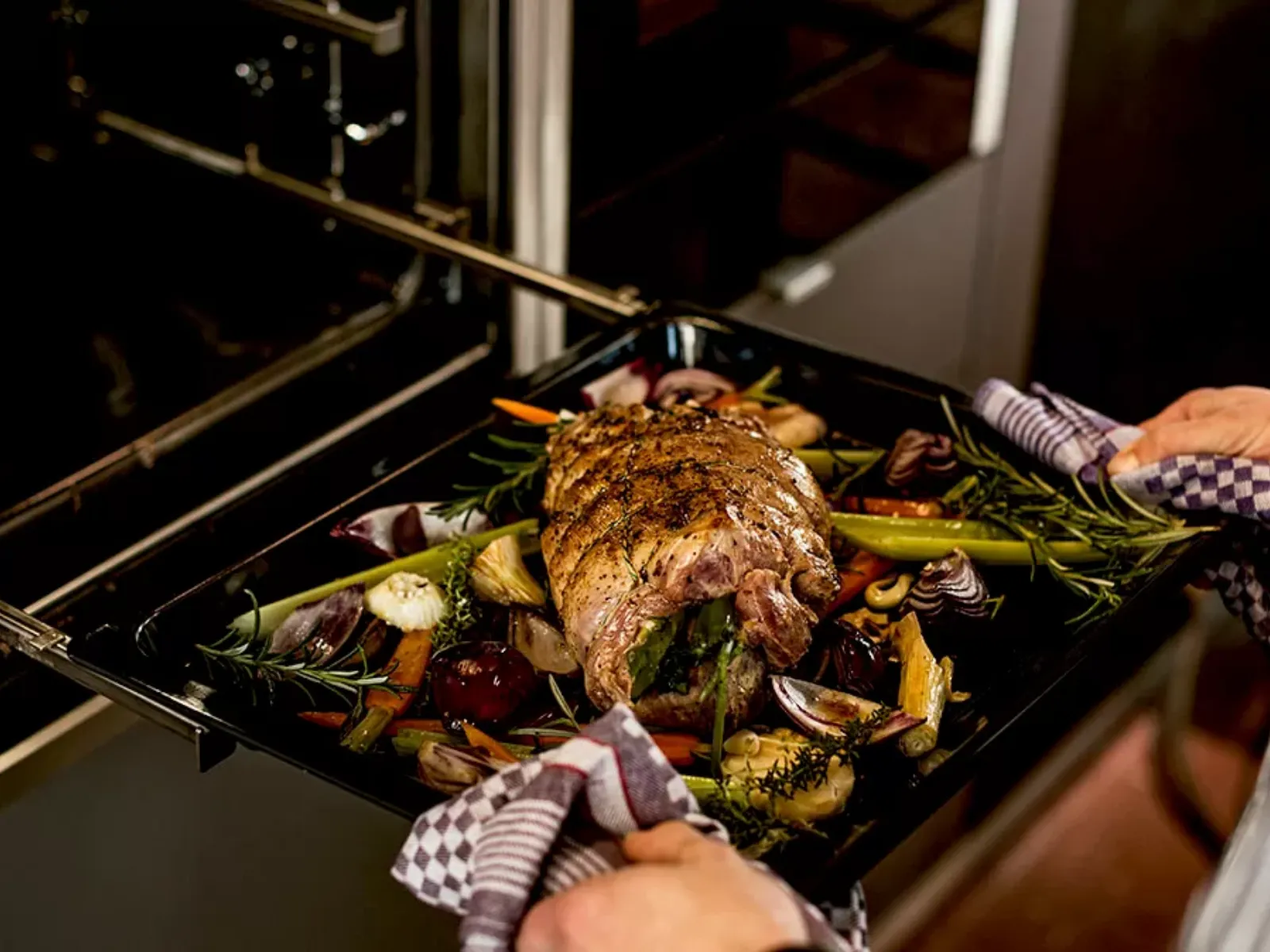 Primo piano di una teglia appena sfornata da un moderno forno da incasso, con un gustoso arrosto ripieno accompagnato da carote arrosto, cipolle rosse, porri ed erbe aromatiche fresche come rosmarino e timo.