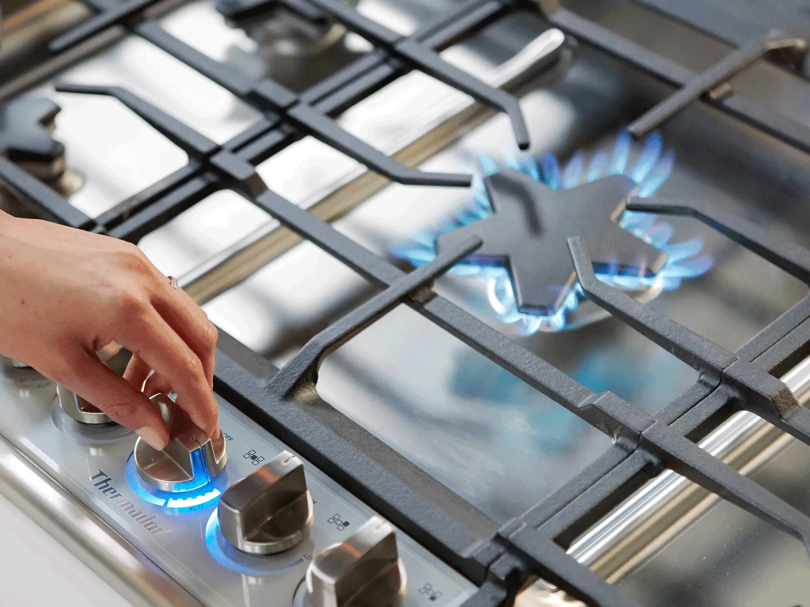 Hand adjusting knob on a Thermador stainless steel gas stove with blue flames under cast iron grates.
