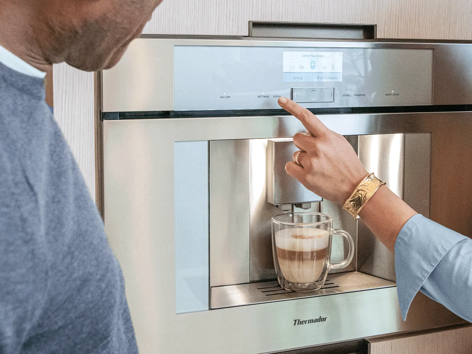 Two people prepare a latte macchiato using a Thermador built-in coffee machine.