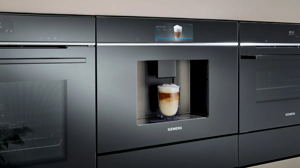 A close-up of a sleek, black Siemens built-in coffee machine dispensing a layered coffee drink into a glass. The machine is flanked by other matching built-in ovens.