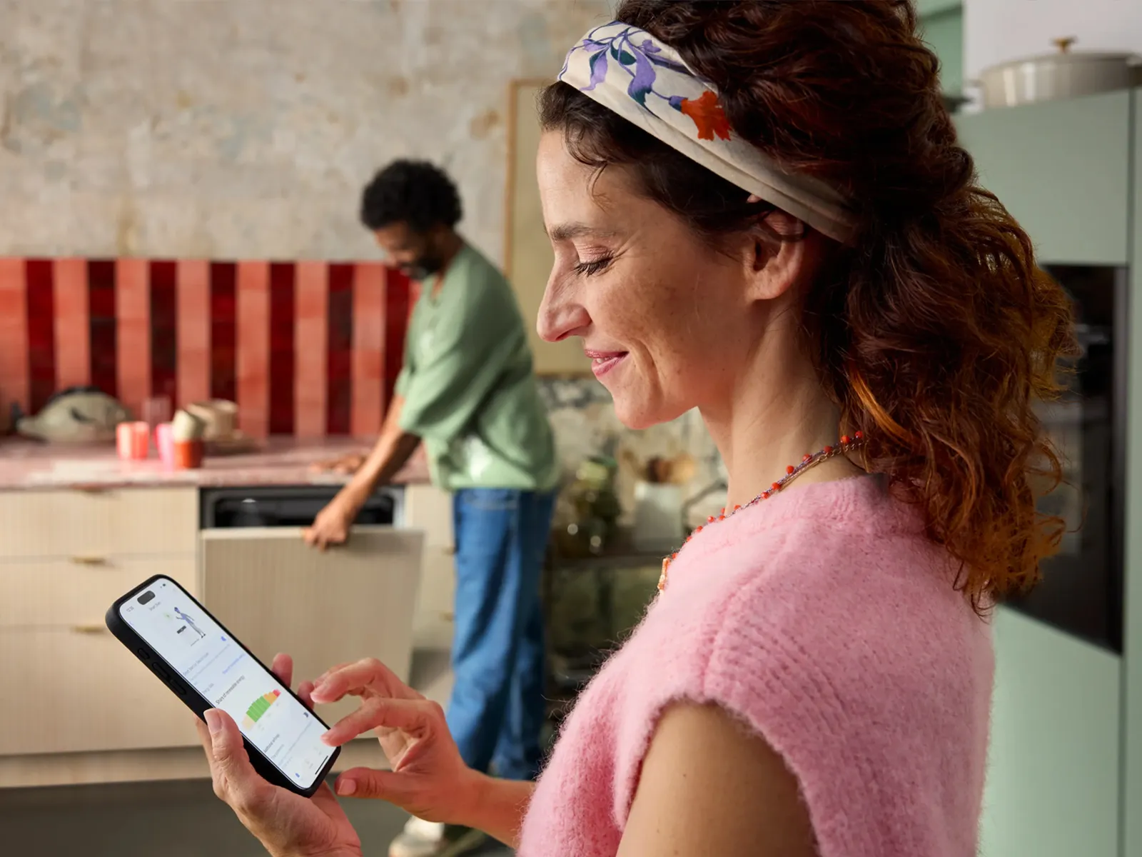 Eine Frau steuert einen NEFF Geschirrspüler über die Home Connect App auf ihrem Smartphone, während ein Mann das Gerät in der Küche belädt.