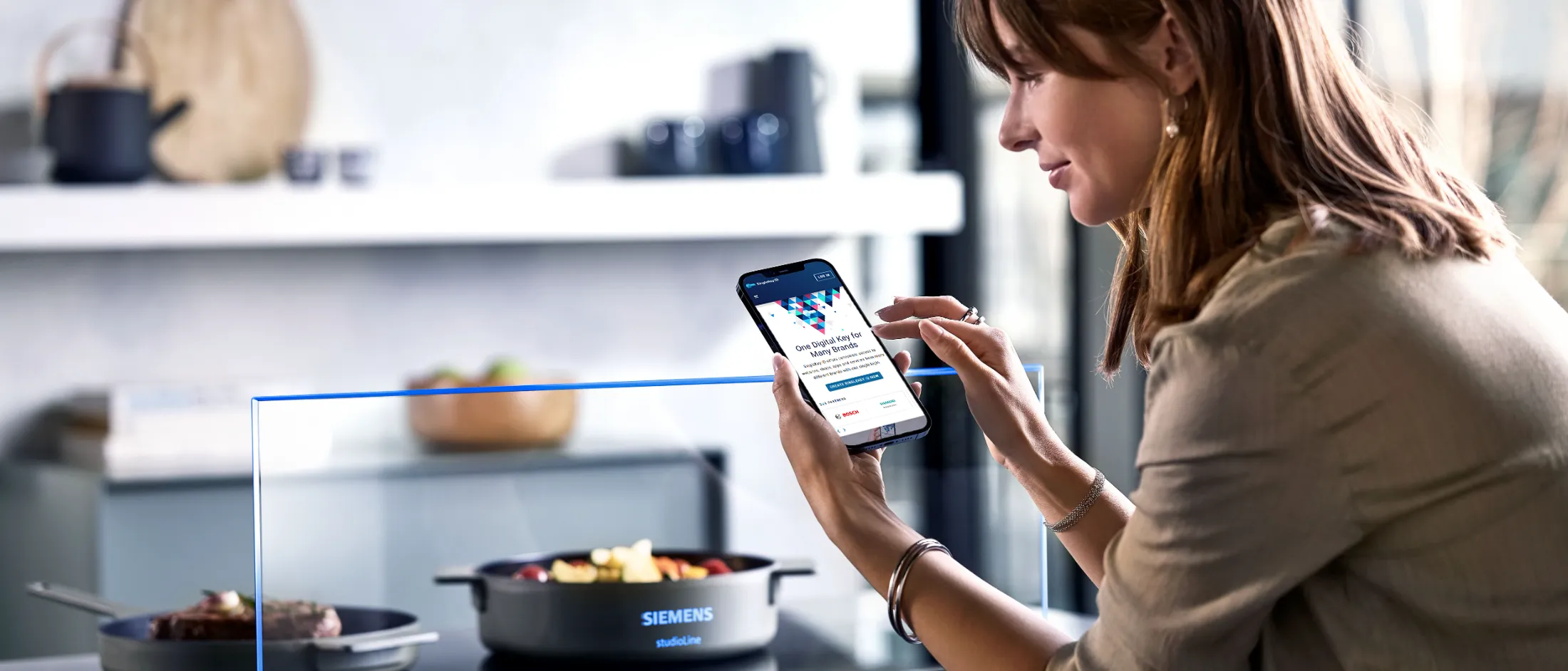 Woman using a smartphone app to control a Siemens studioLine kitchen appliance with food cooking inside.
