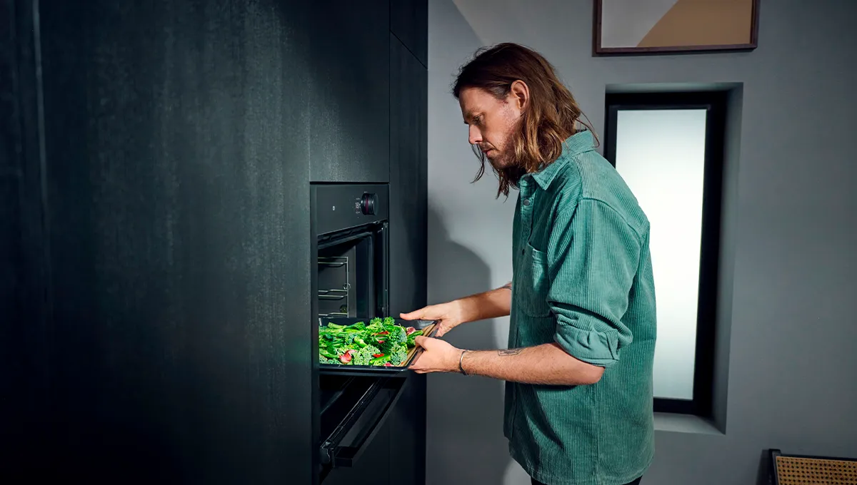 Uomo con camicia color foglia di tè che mette una teglia di verdure fresche in un forno nero moderno in una cucina minimalista.