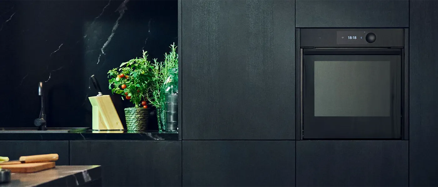 Interno di una cucina scura moderna con forno da incasso, rubinetto nero, piante aromatiche e un ceppo di coltelli sul bancone.