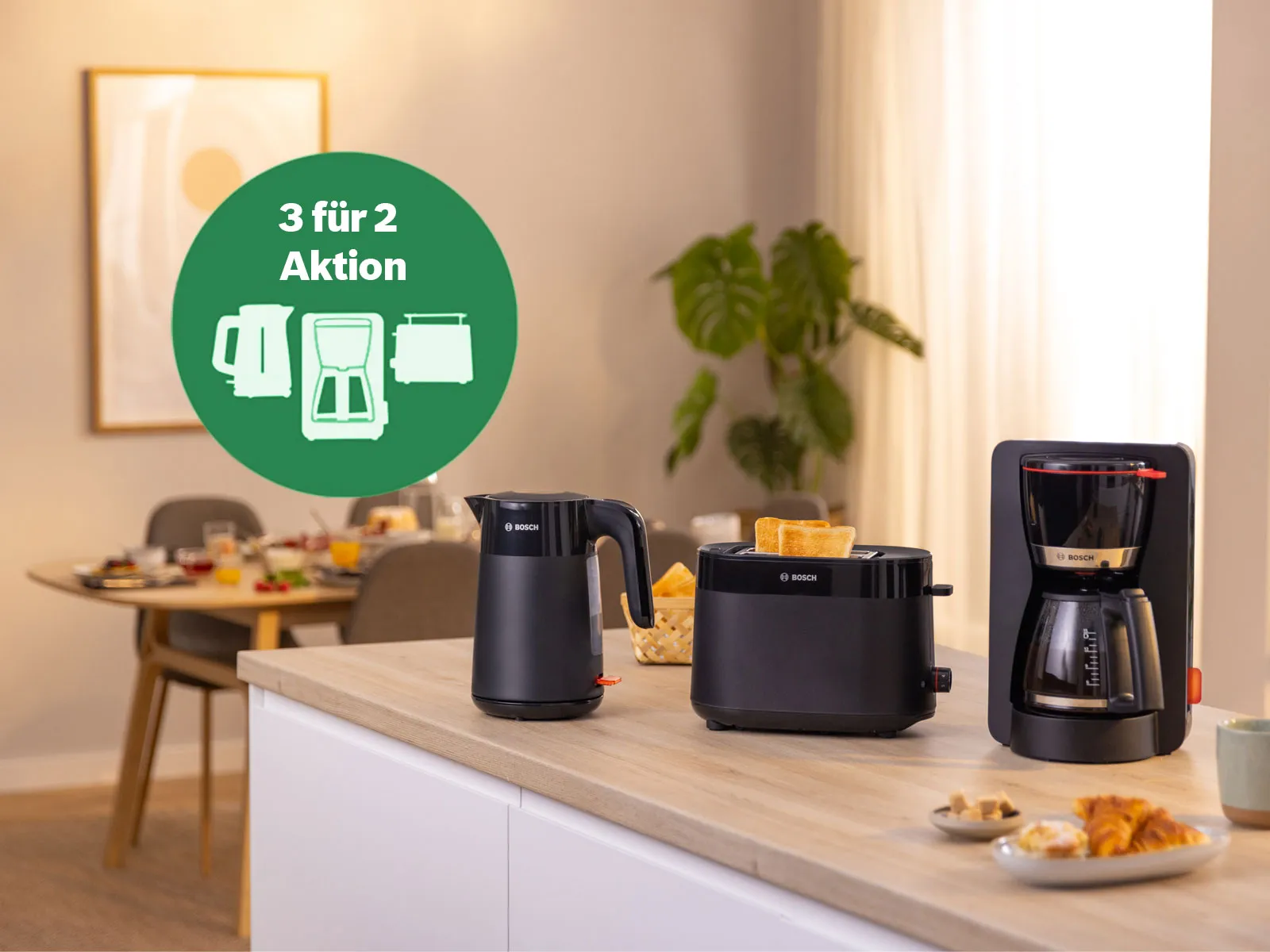 Auf einem Küchenblock werden Bosch Frühstücksgeräte bestehend aus einem schwarzen Wasserkocher, Toaster und einer Filterkaffeemaschine mit dem Hinweis auf die aktuelle Bosch 3für2 Aktion gezeigt.