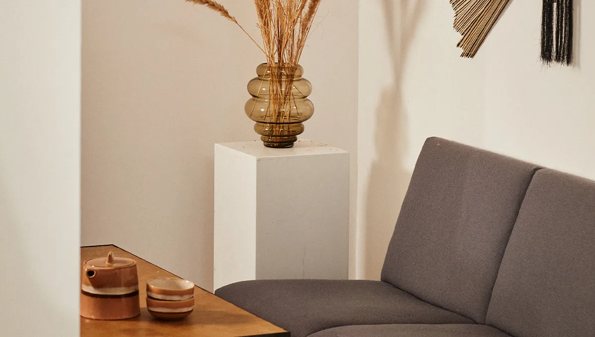 Un soggiorno minimalista in stile Japandi con un divano grigio, un tavolino in legno con una teiera in ceramica e un vaso di erba della pampa essiccata su un piedistallo bianco.