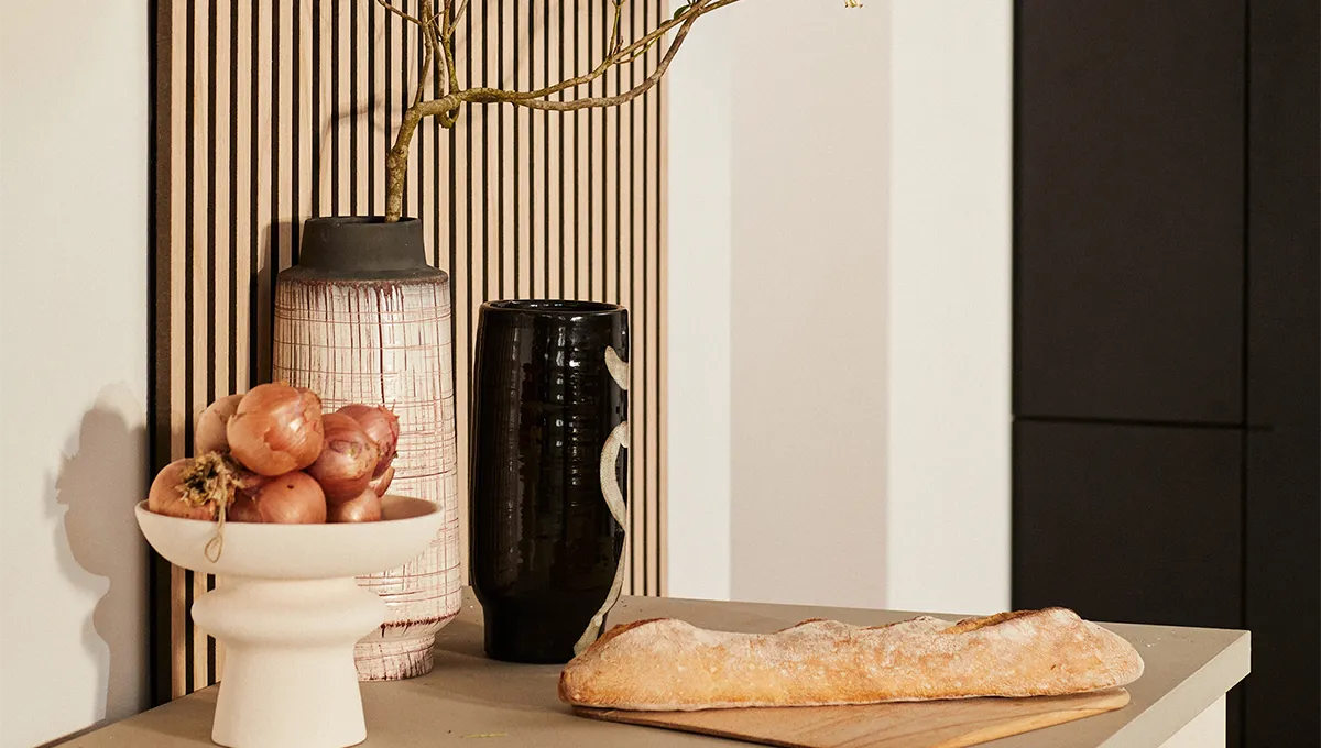 Un primo piano di un bancone da cucina in stile Japandi, che mostra una baguette rustica, una ciotola di scalogni e vasi artigianali disposti di fronte a un pannello a parete a doghe di legno chiaro.