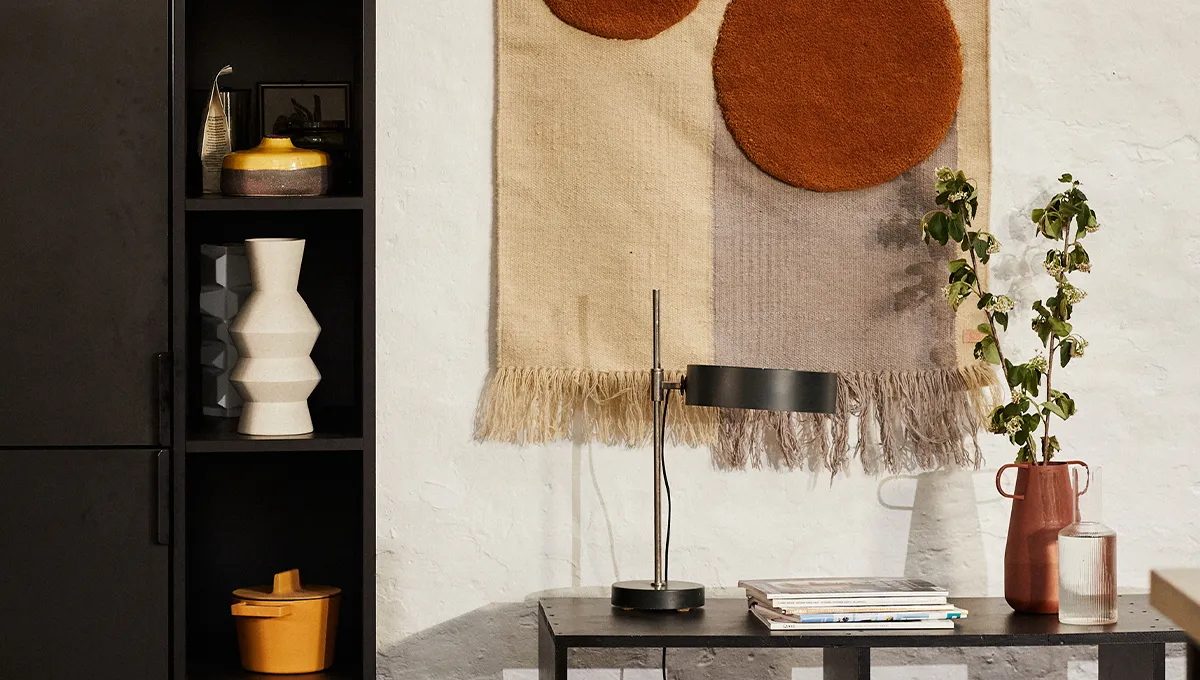 Un angolo accogliente decorato in stile Japandi, con un tavolino nero con una lampada, mensole nere con ceramiche e un grande arazzo strutturato nei toni del neutro e del ruggine.