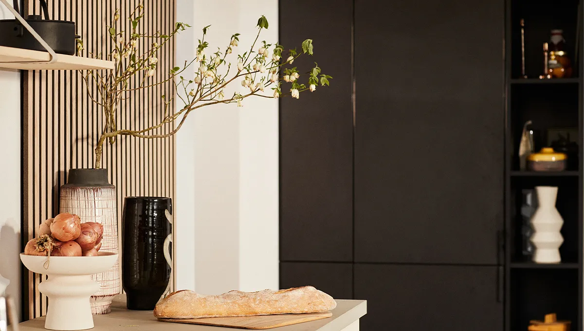 Una vignetta di cucina in stile Japandi con una baguette fresca, una ciotola di scalogni e vasi decorativi su uno sfondo di una parete a doghe di legno e mobili neri minimalisti.