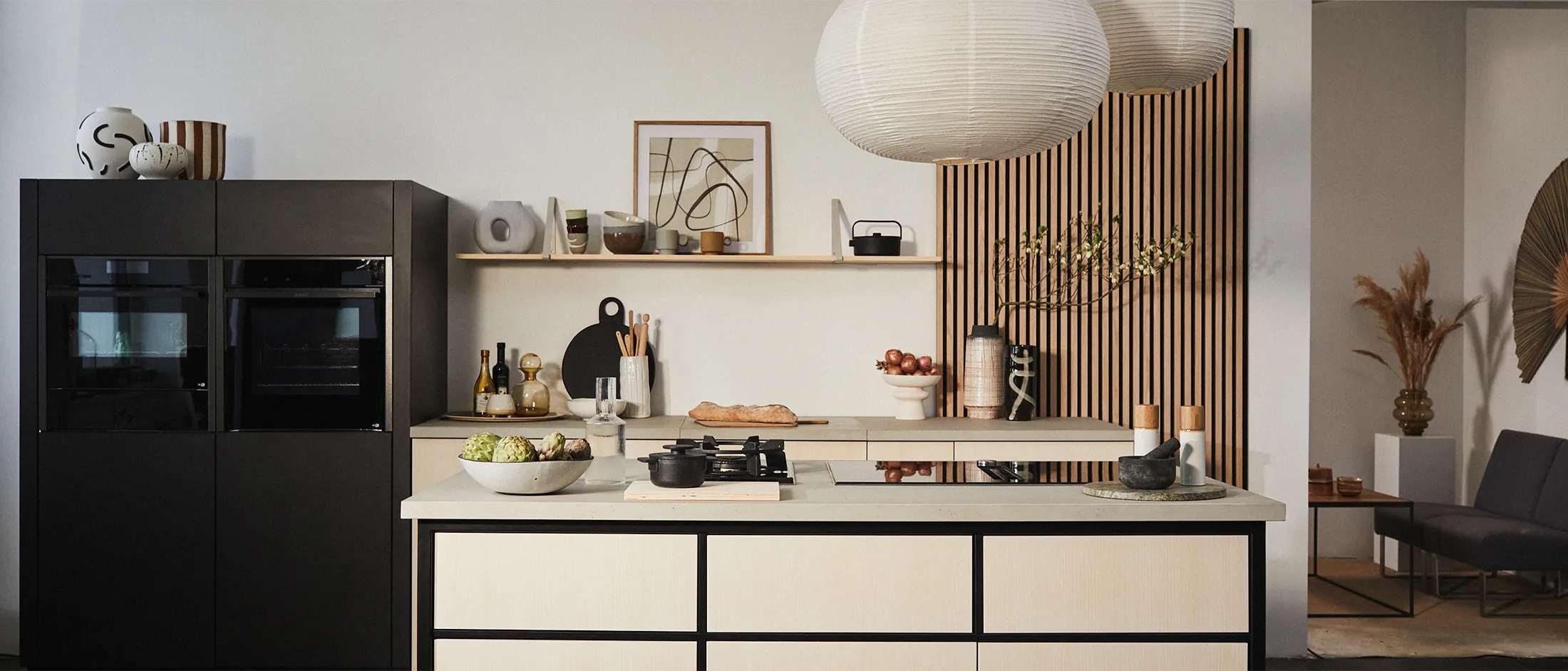 Un'ampia inquadratura di una cucina completa in stile Japandi, con forni neri NEFF, un'isola in legno chiaro con un piano cottura a domino e un arredamento minimalista che include grandi lanterne di carta e mensole aperte.