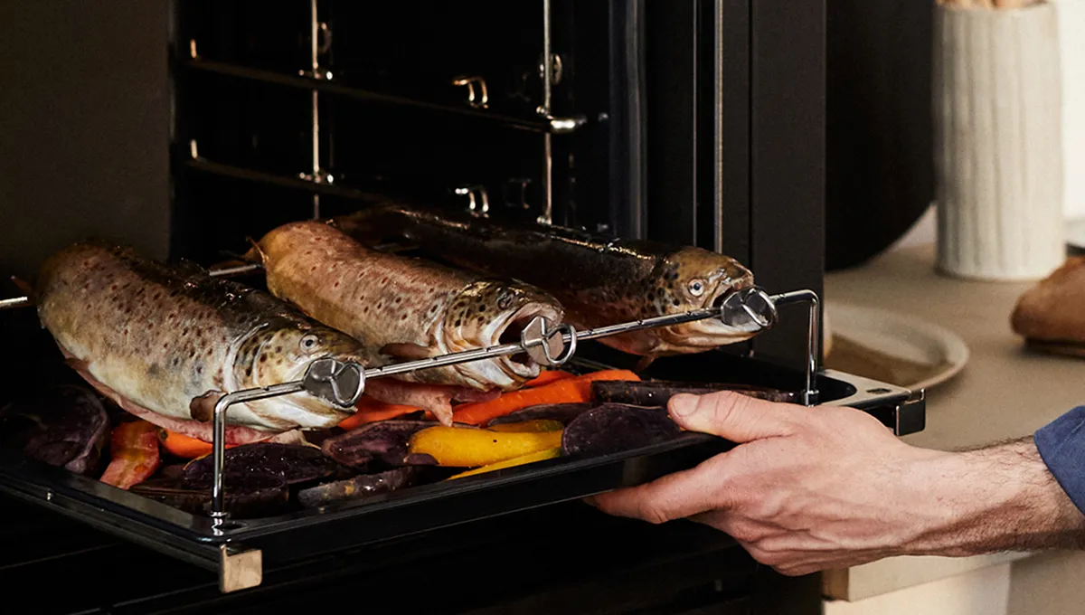 Un primo piano di una mano che estrae una teglia da un forno NEFF, mostrando tre pesci interi che arrostiscono sull'accessorio girarrosto sopra un letto di ortaggi a radice.