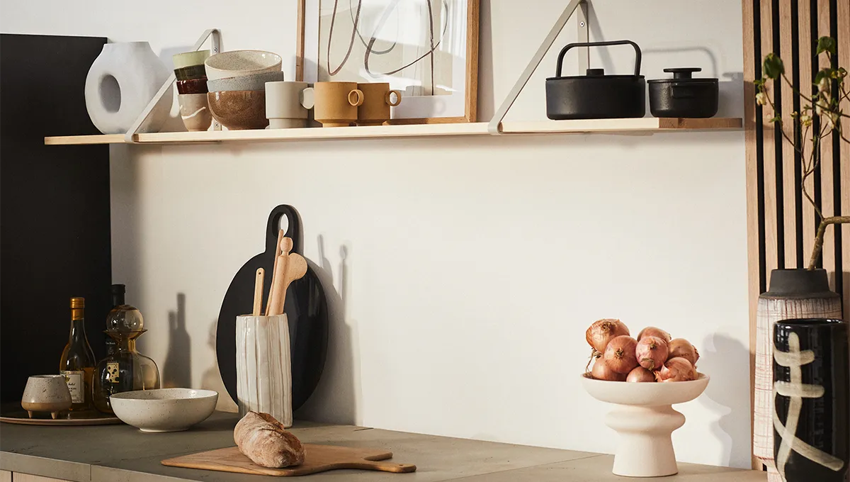 Un bancone da cucina minimalista in stile Japandi con una mensola aperta in legno chiaro che contiene ceramiche e arte astratta, sopra una collezione di taglieri, ciotole e una pagnotta di pane fresco.