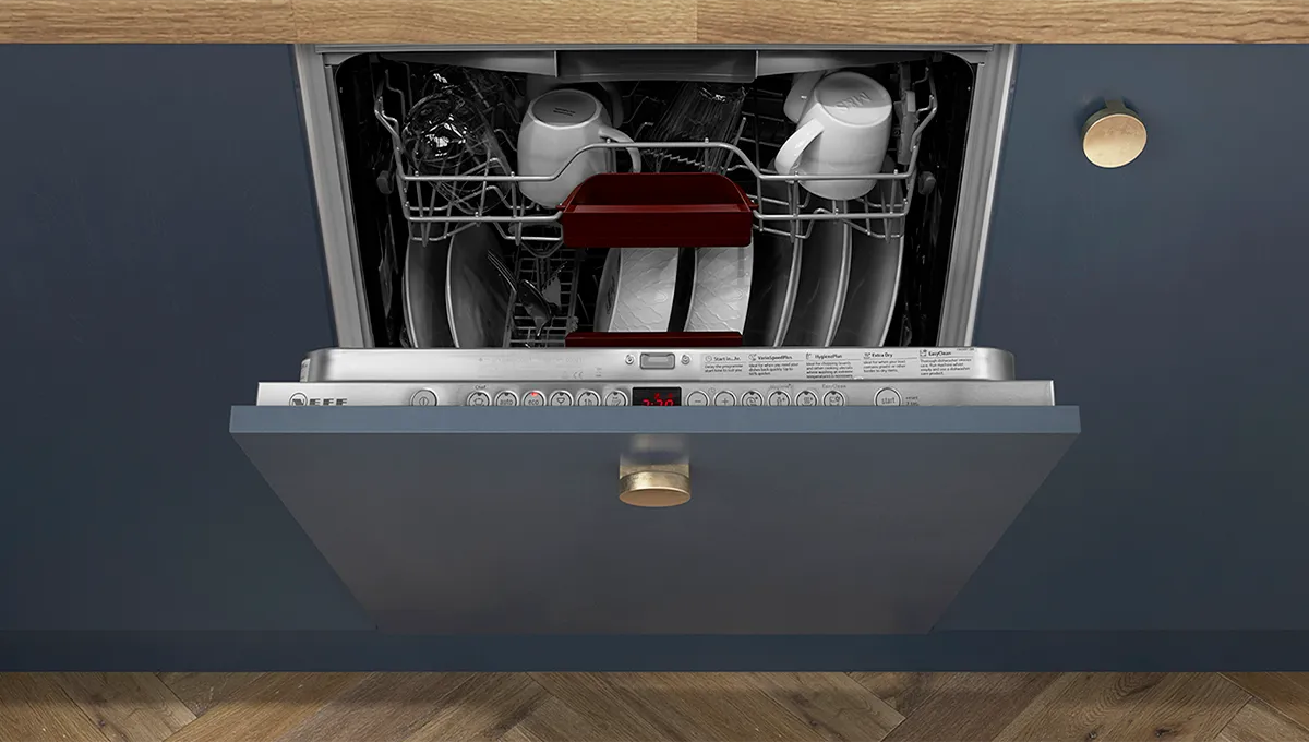 A top-down view of an open, fully integrated NEFF dishwasher with a blue cabinet front, showcasing a loaded top rack with clean white mugs and plates.