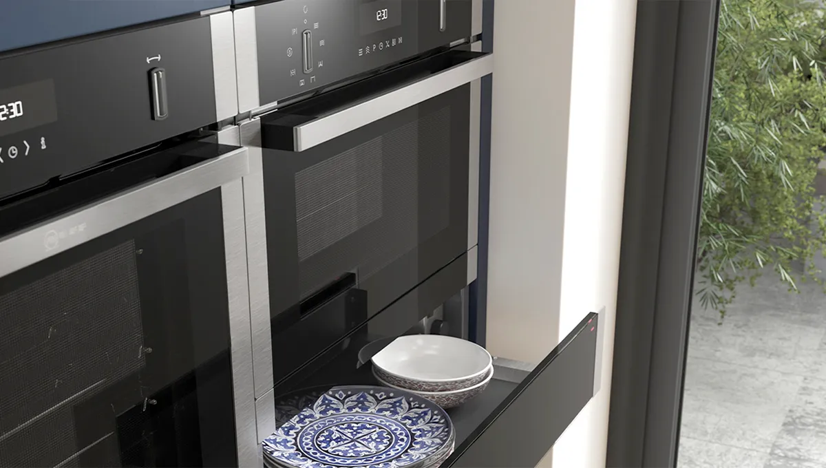 A close-up shot of an open NEFF warming drawer, positioned beneath a stack of built-in ovens and filled with elegant blue and white patterned plates and bowls.