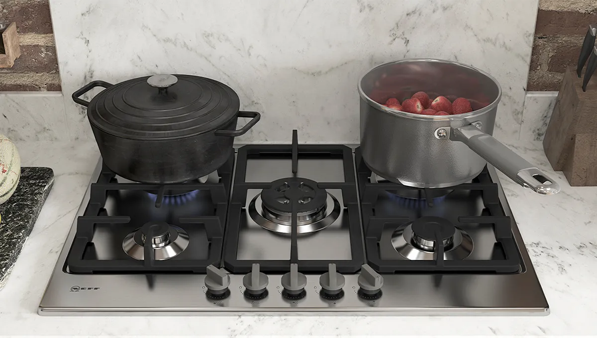A close-up of a stainless steel NEFF gas hob in action, with a black cast-iron pot and a saucepan of strawberries cooking on the burners against a white marble backsplash.